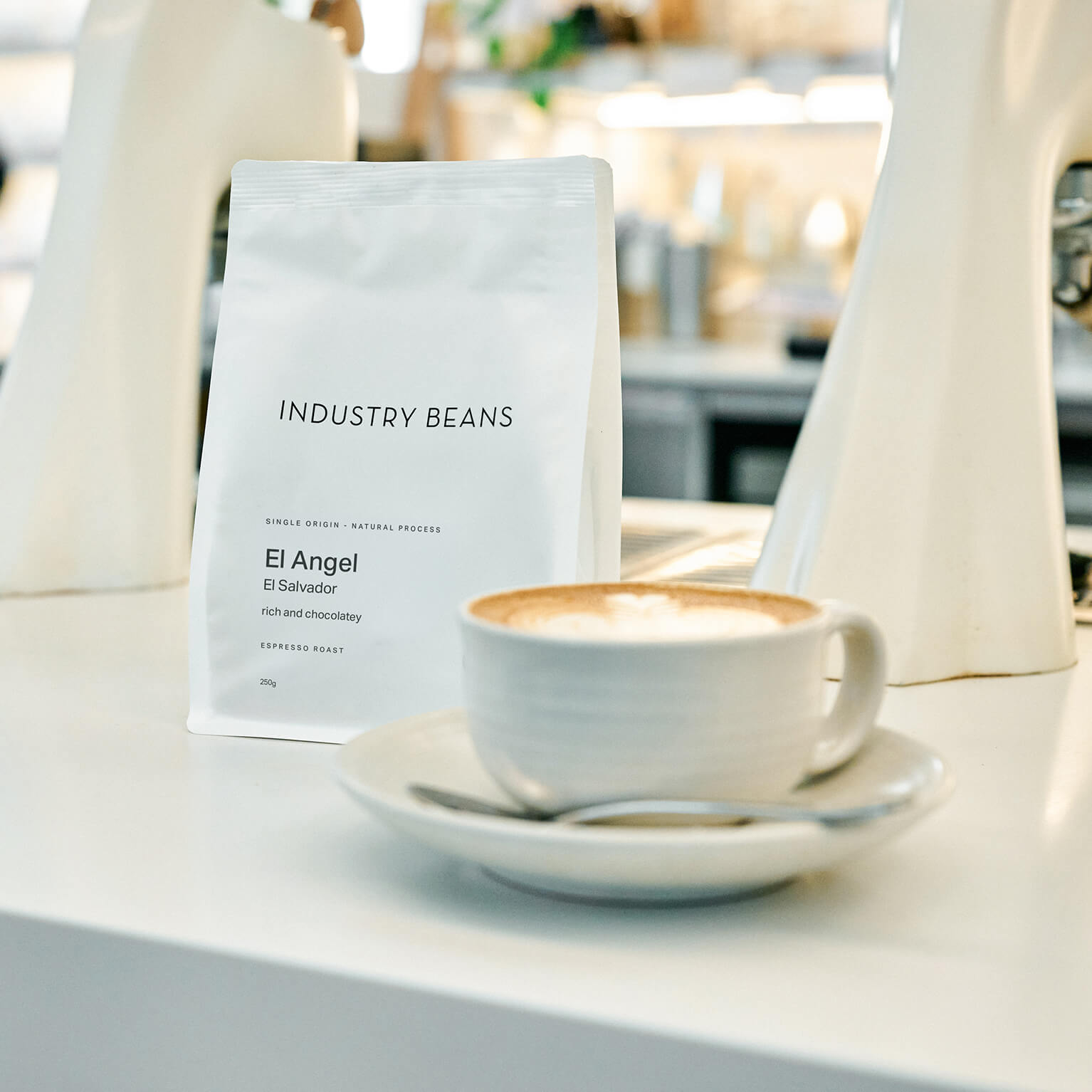 A white bag of Industry Beans El Angel espresso roast coffee stands on a white counter next to a white ceramic cup of cappuccino with latte art, with a blurred cafe interior in the background.