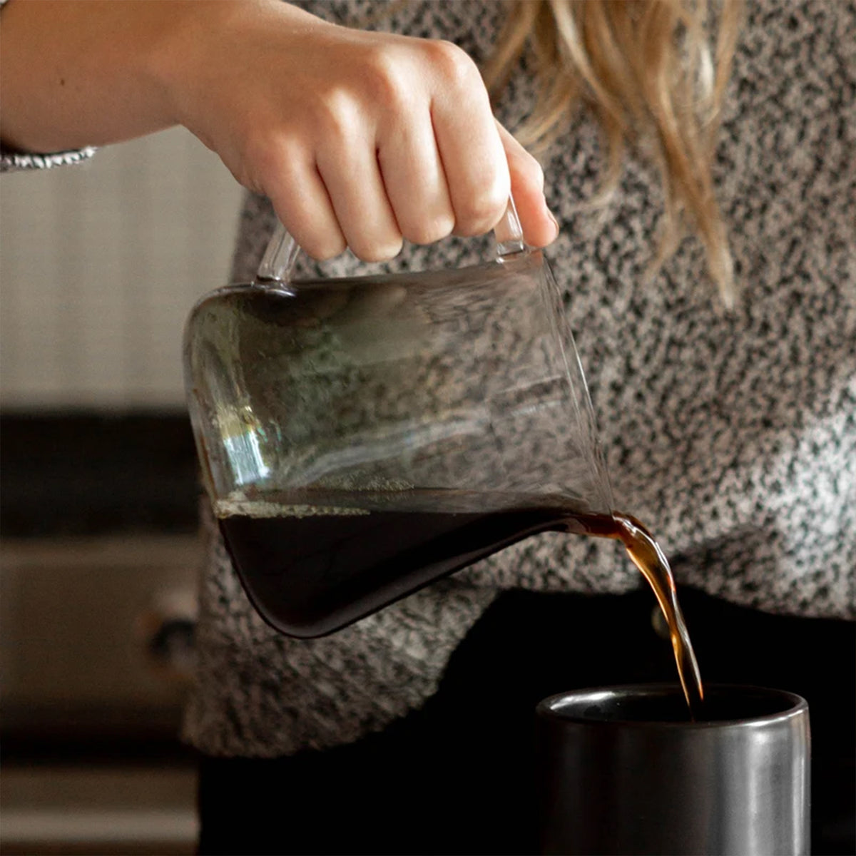 Hand pouring brewed coffee from the Fellow Mighty Small Glass Carafe into a dark ceramic mug