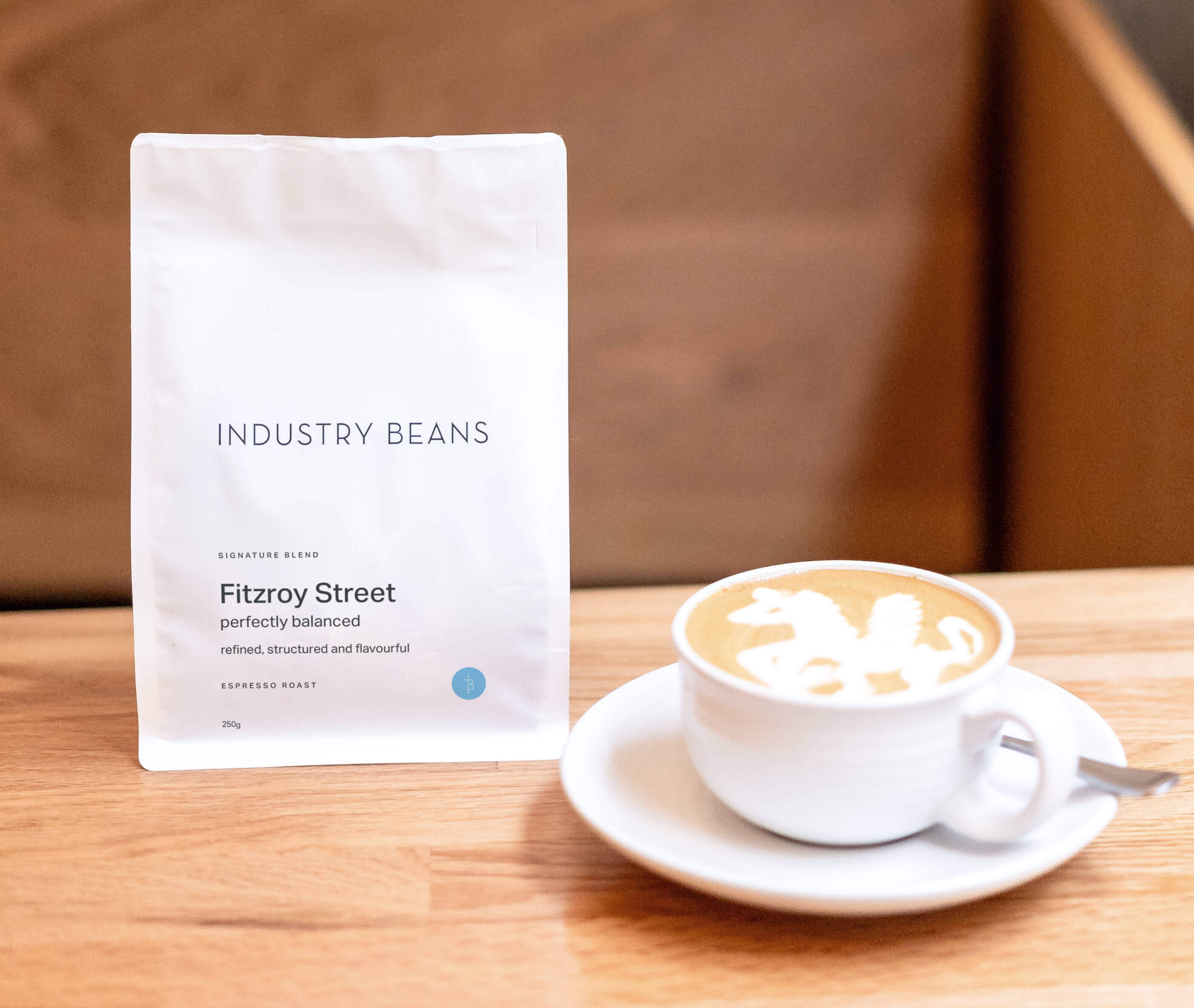 Coffee cup with latte art next to a package of 'Industry Beans Fitzroy Street' on a wooden surface.