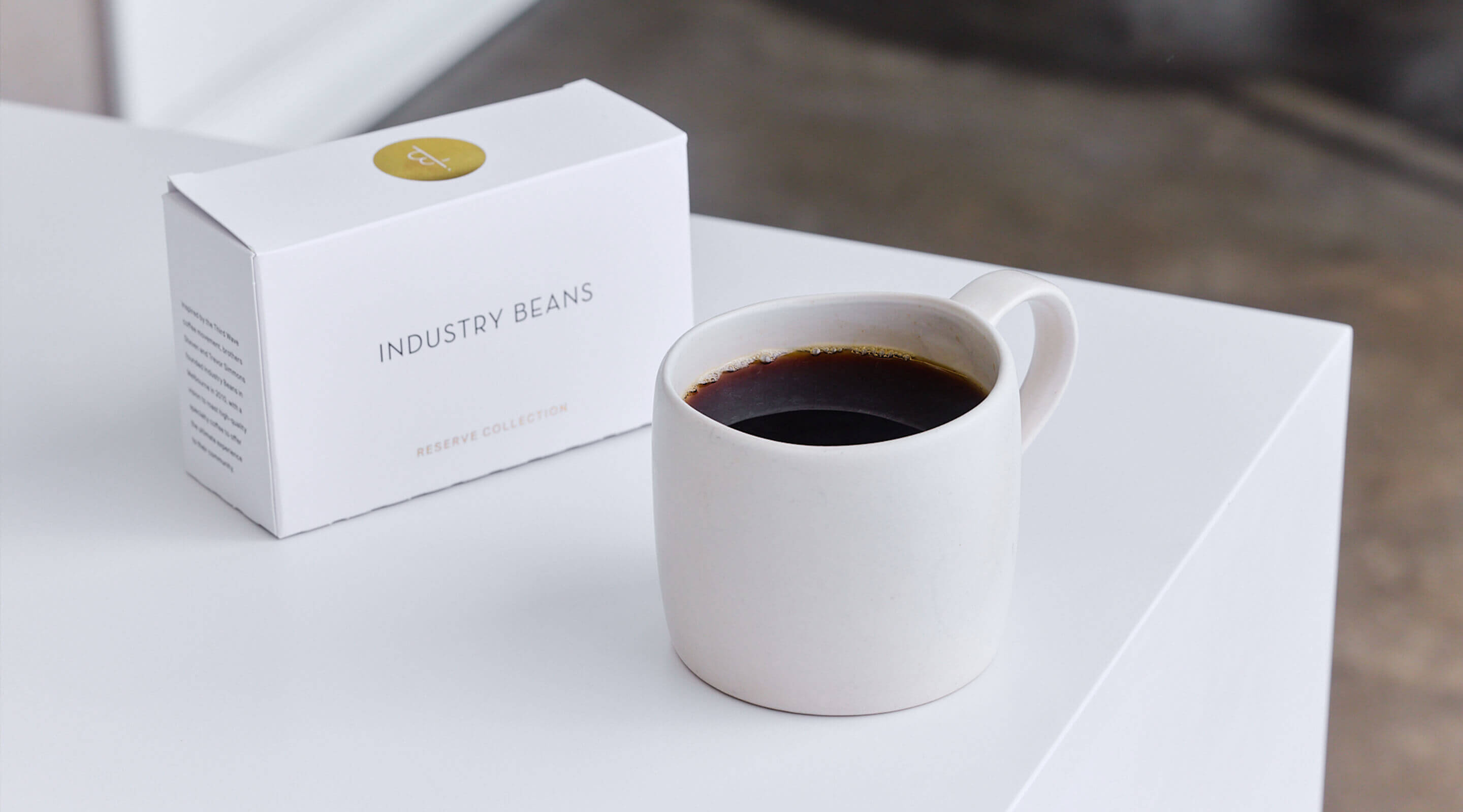 White mug with coffee and 'Industry Beans' packaging on a white surface
