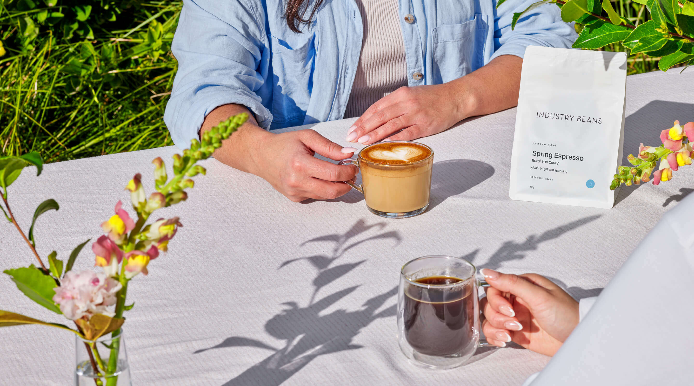 Two people sitting at a table outdoors with coffee and a bag of coffee beans.
