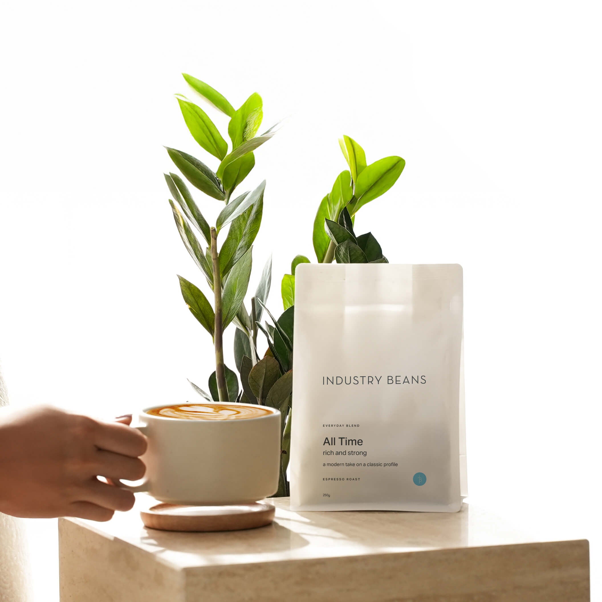 A white bag of Industry Beans coffee stands on a light wood block next to a hand holding a cup of coffee, with green plants in the background against a bright white wall.
