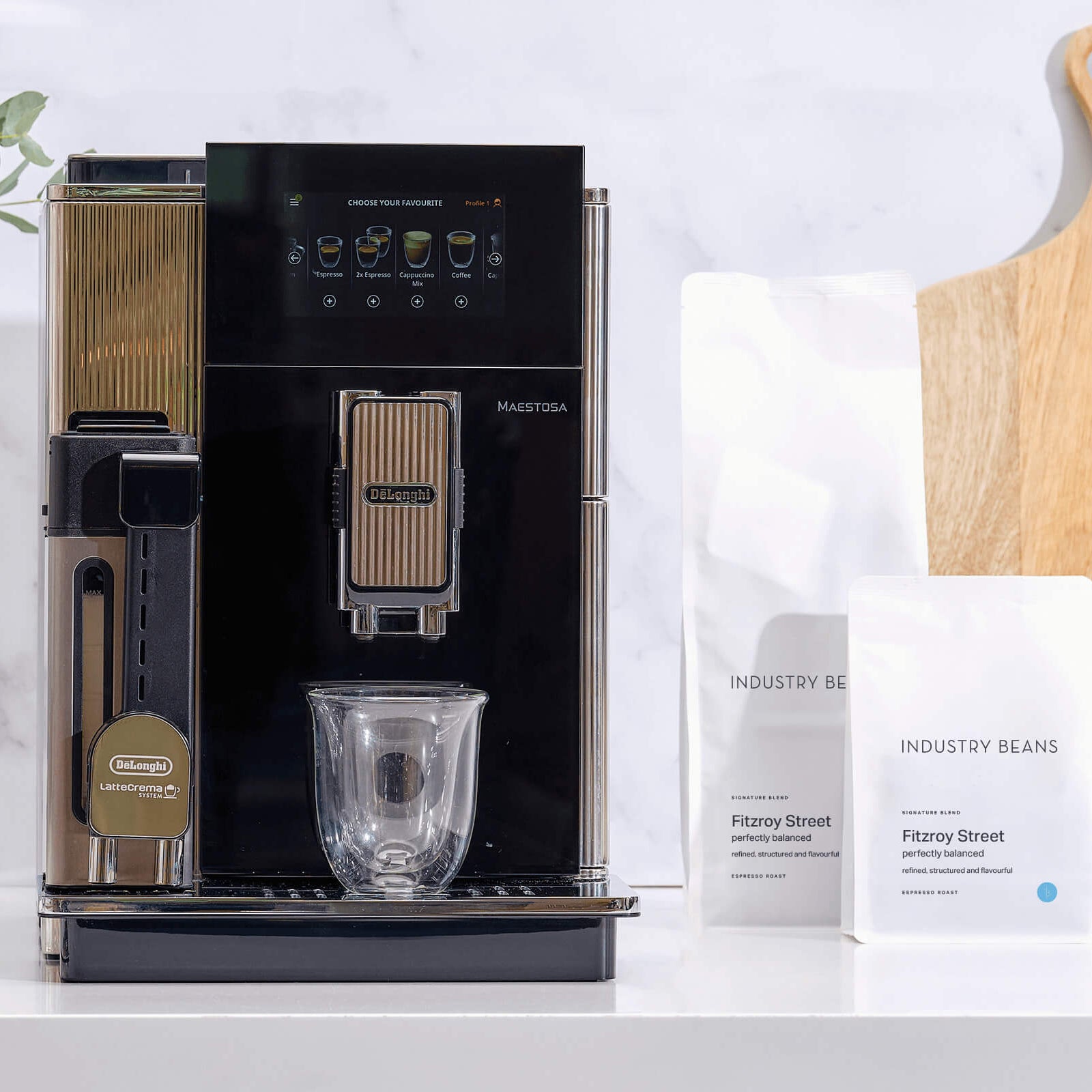 Black coffee machine on a white countertop with a plant and bags of coffee beans in the background.