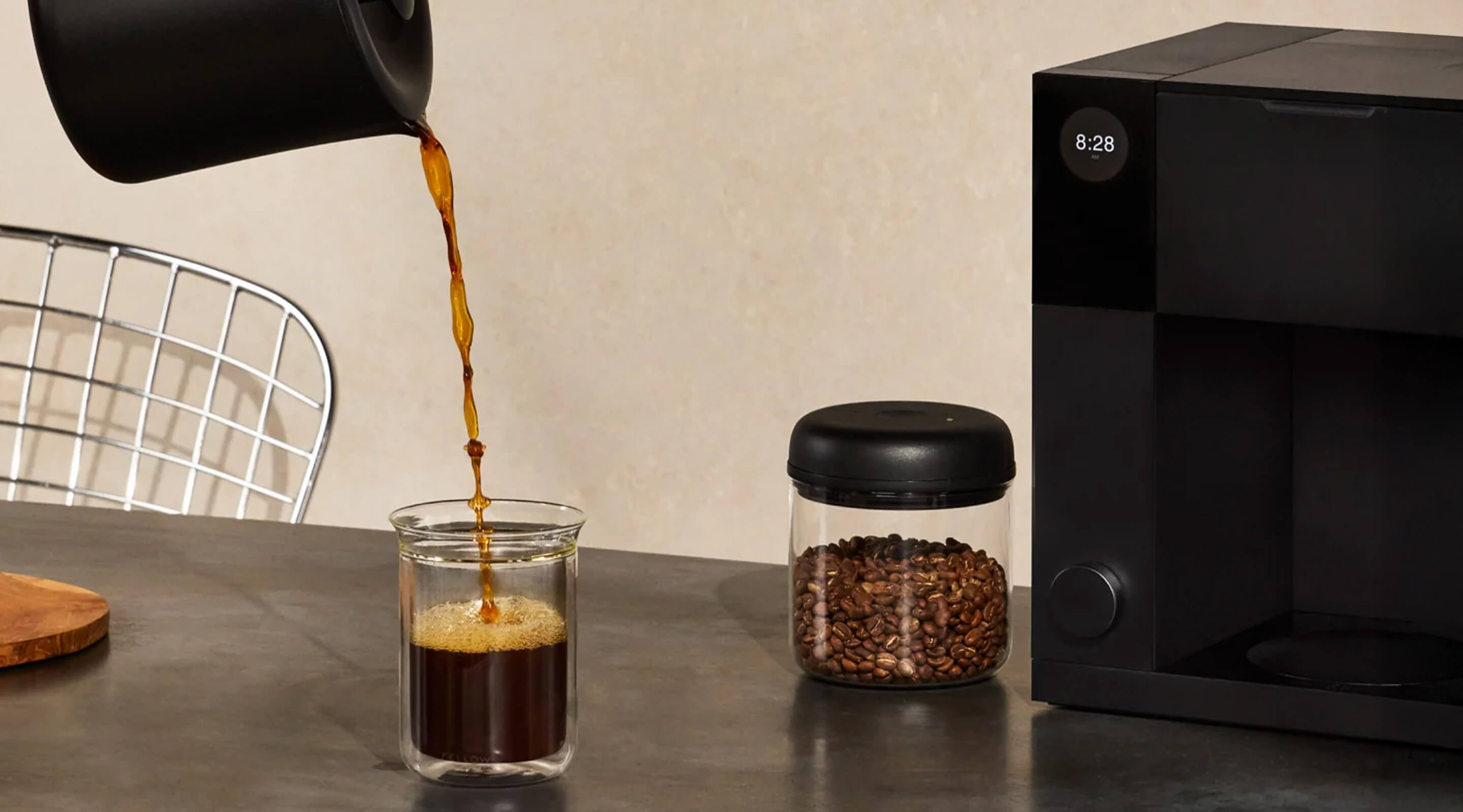 Coffee being poured into a glass, with coffee beans and a coffee maker in the background.