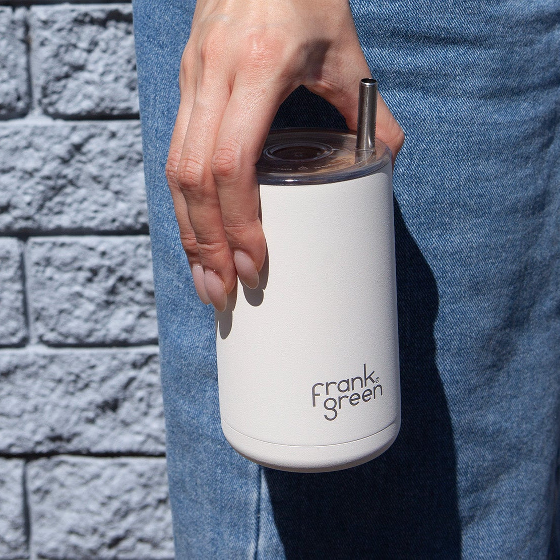 Person holding a white reusable coffee cup with 'Frank Green' branding against a brick wall.