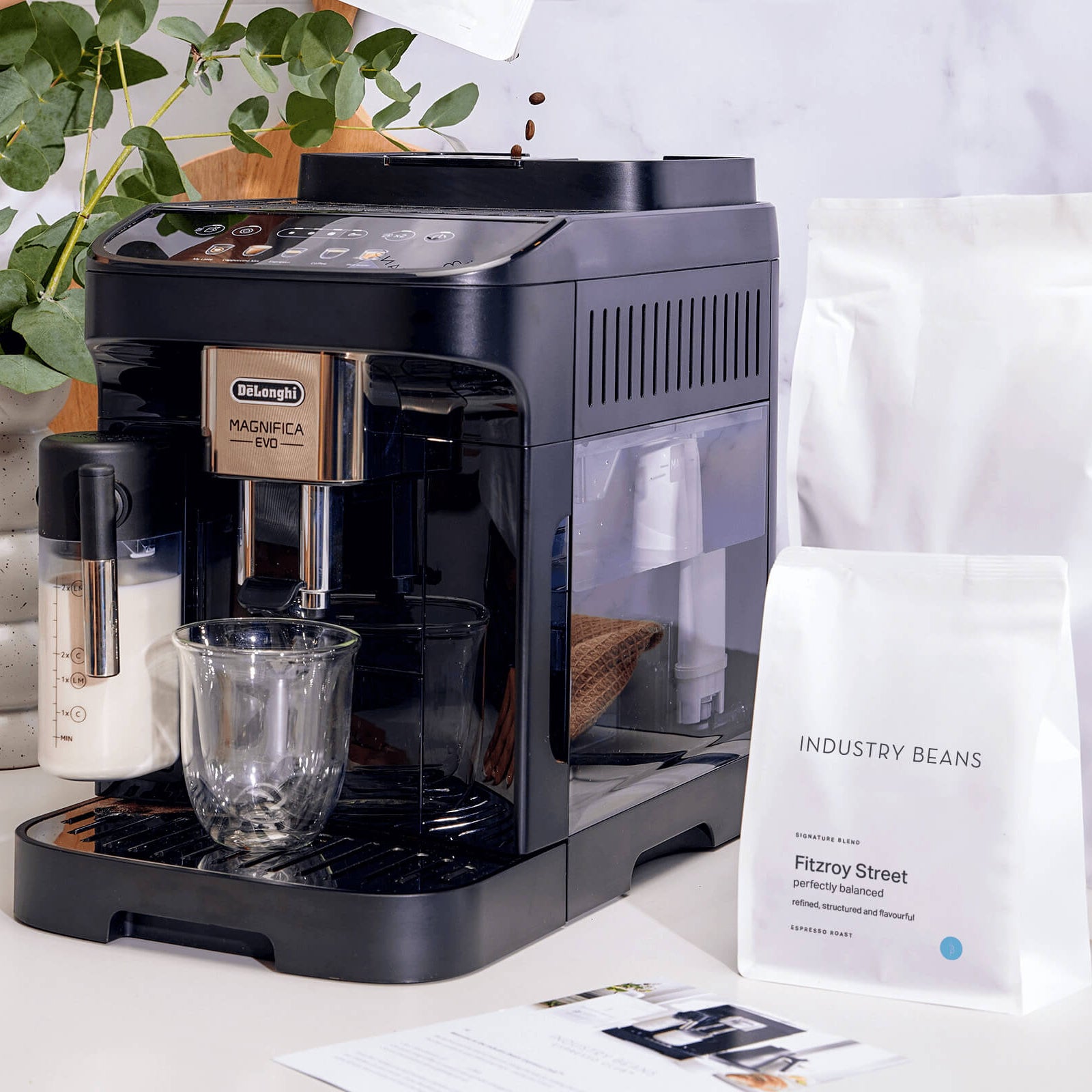 Black coffee machine on a white countertop with coffee bags and a plant in the background.