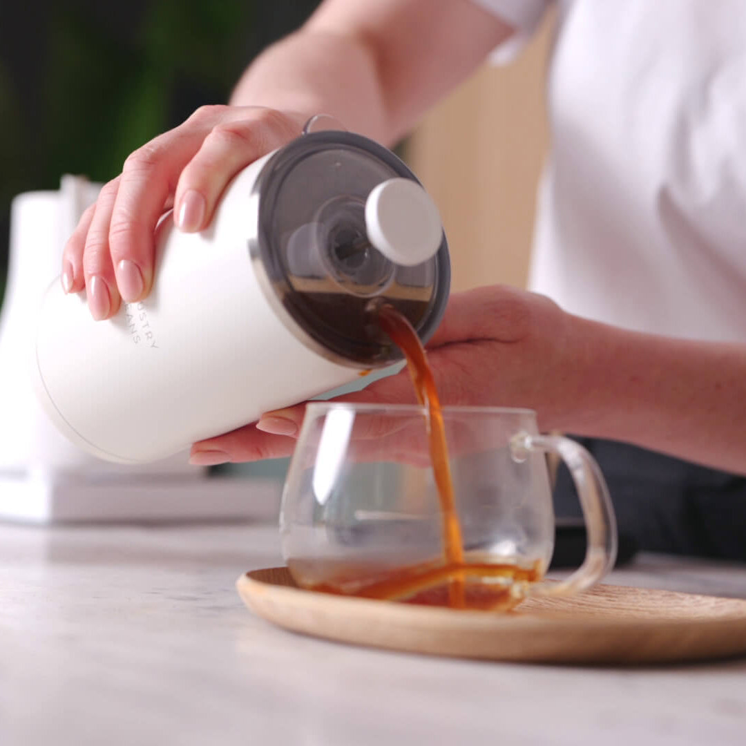 Person pouring coffee from a white container into a glass mug on a wooden coaster.