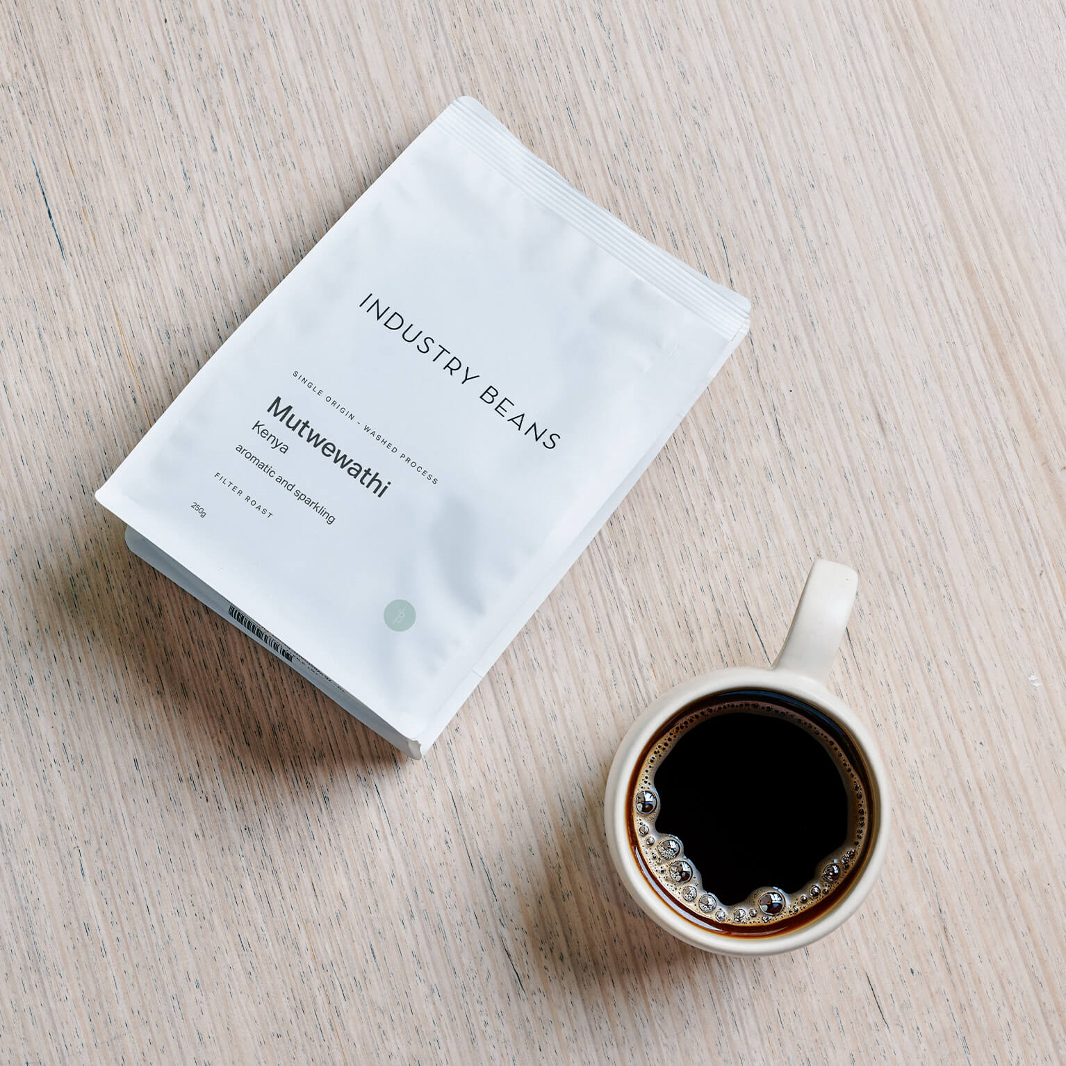 A white bag of Industry Beans Mutwewathi coffee beans lies on a light wood surface next to a white ceramic cup filled with black coffee.