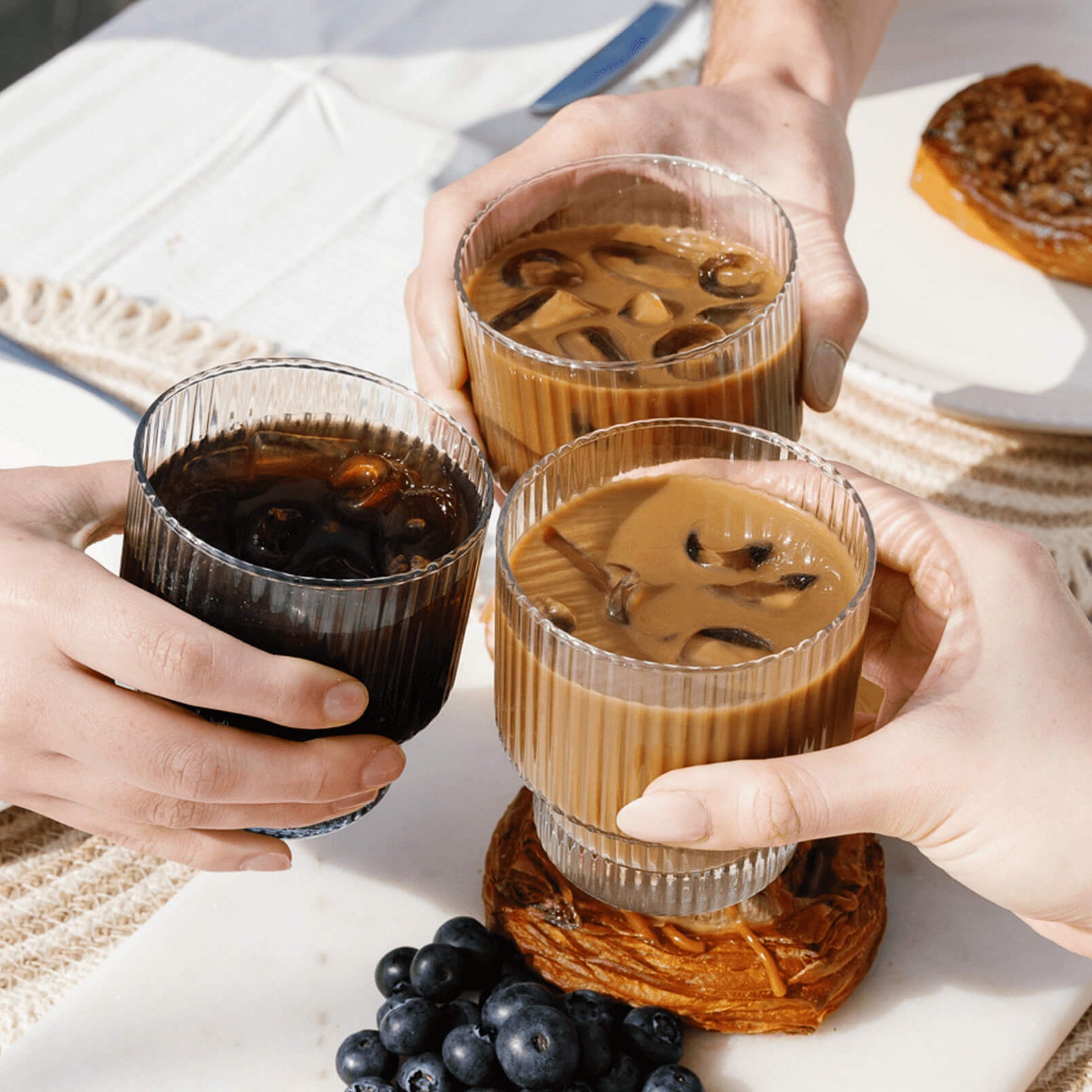 Four hands holding glasses of iced coffee on a table with pastries and grapes.