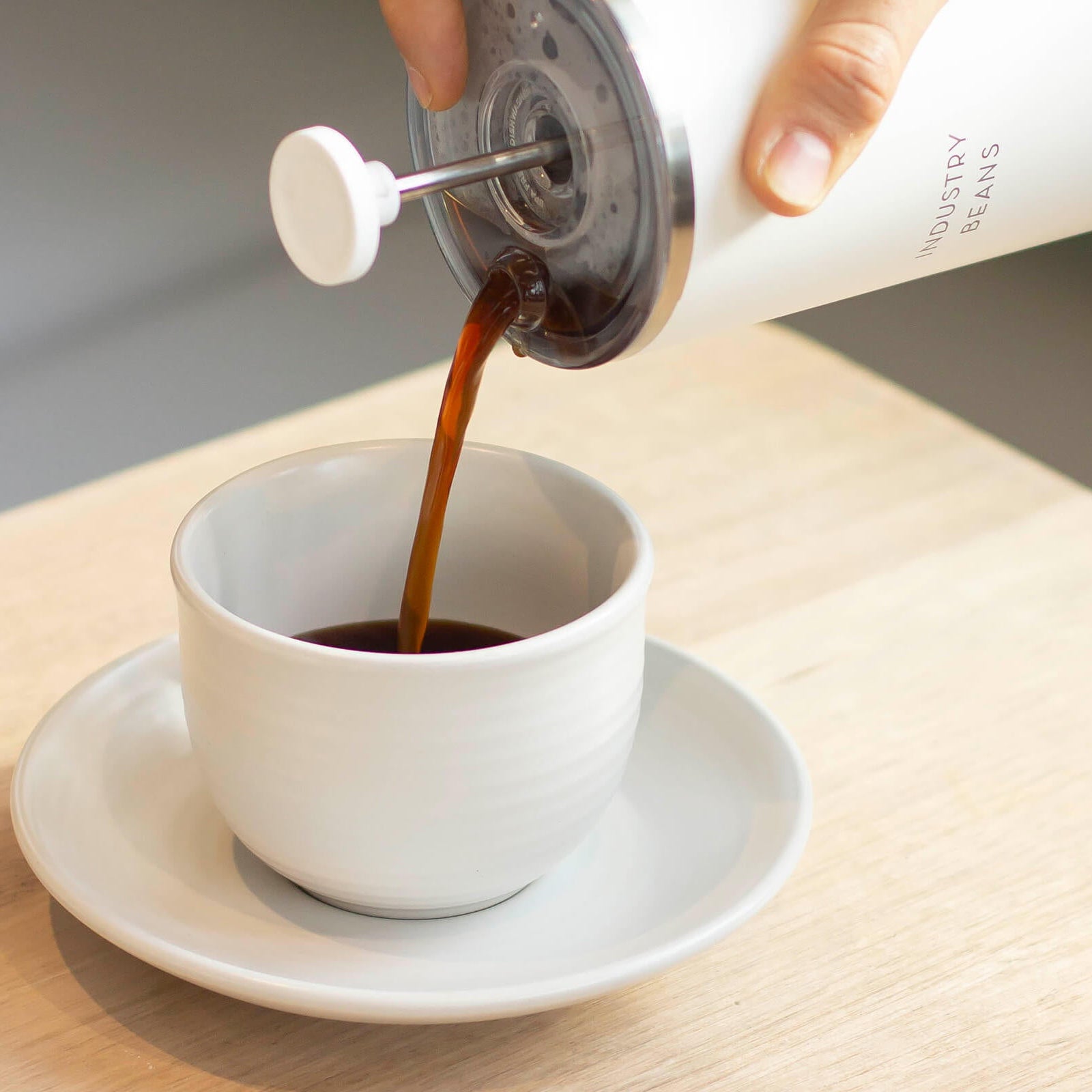 Person pouring coffee from a white container into a white cup on a wooden table.