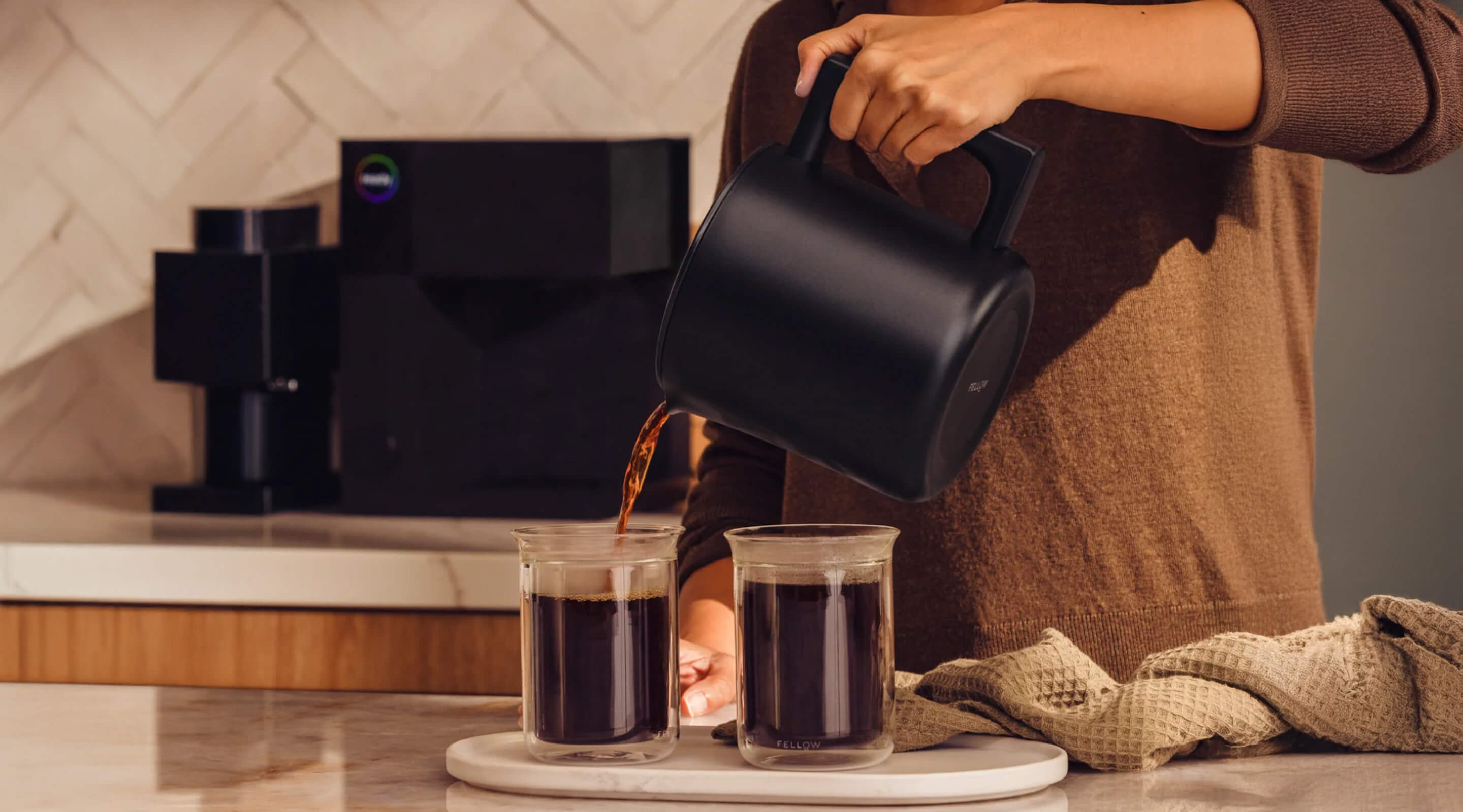Person pouring coffee from a black coffee maker into two glasses on a kitchen counter.