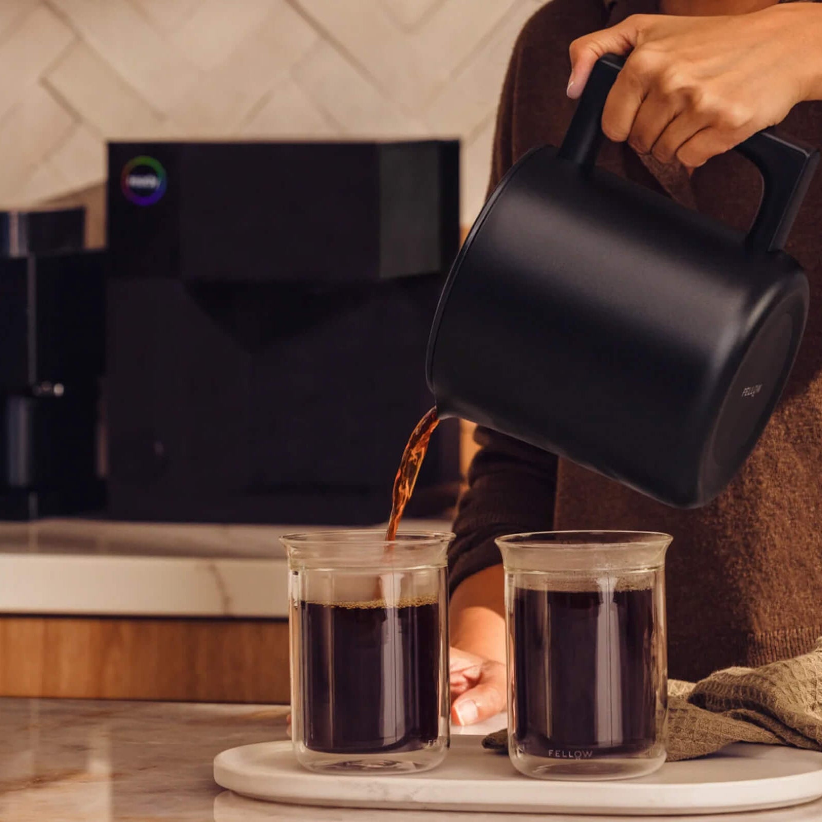 Person pouring coffee from a black coffee maker into two glasses on a kitchen counter.
