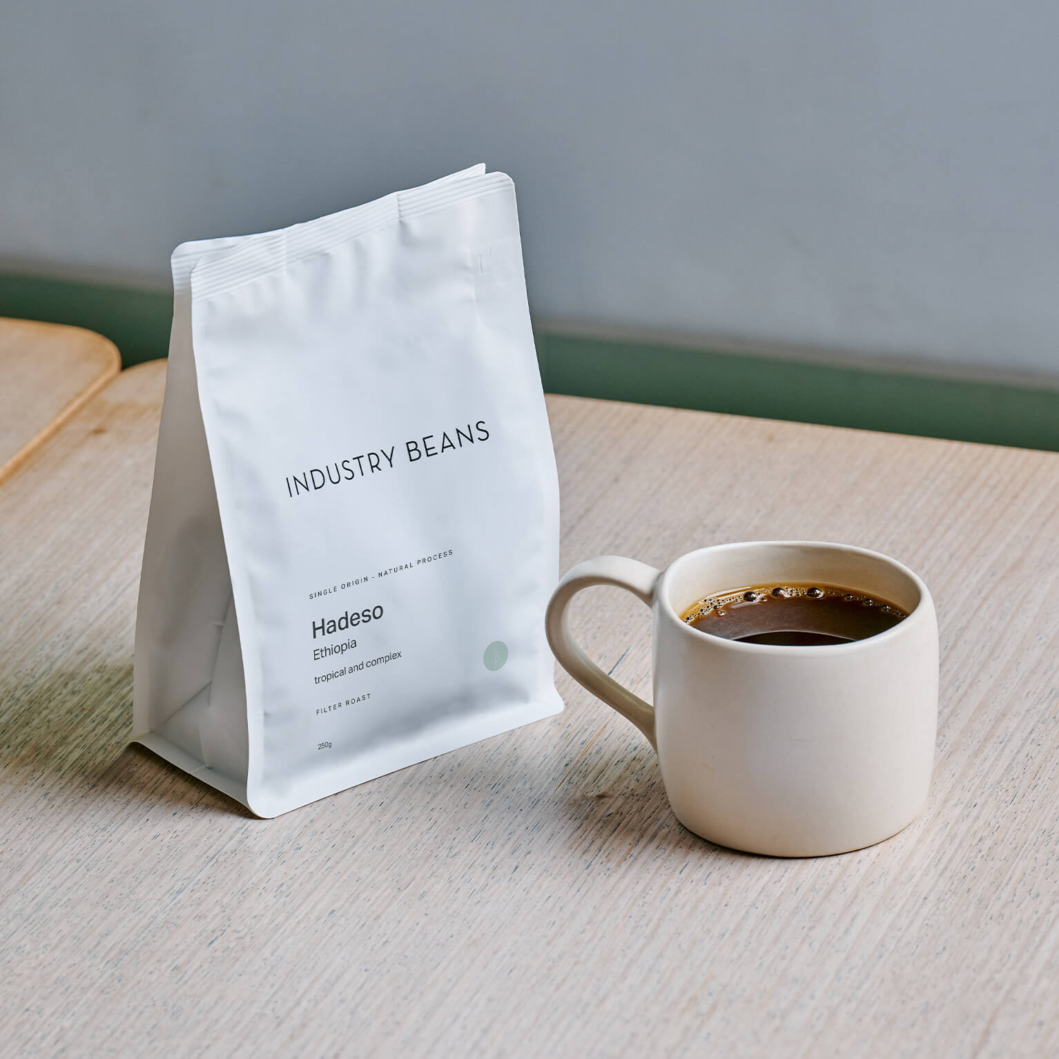 A white bag of Industry Beans Hadeso single origin coffee sits on a light wood table next to a matching ceramic mug filled with black coffee, against a neutral gray background.