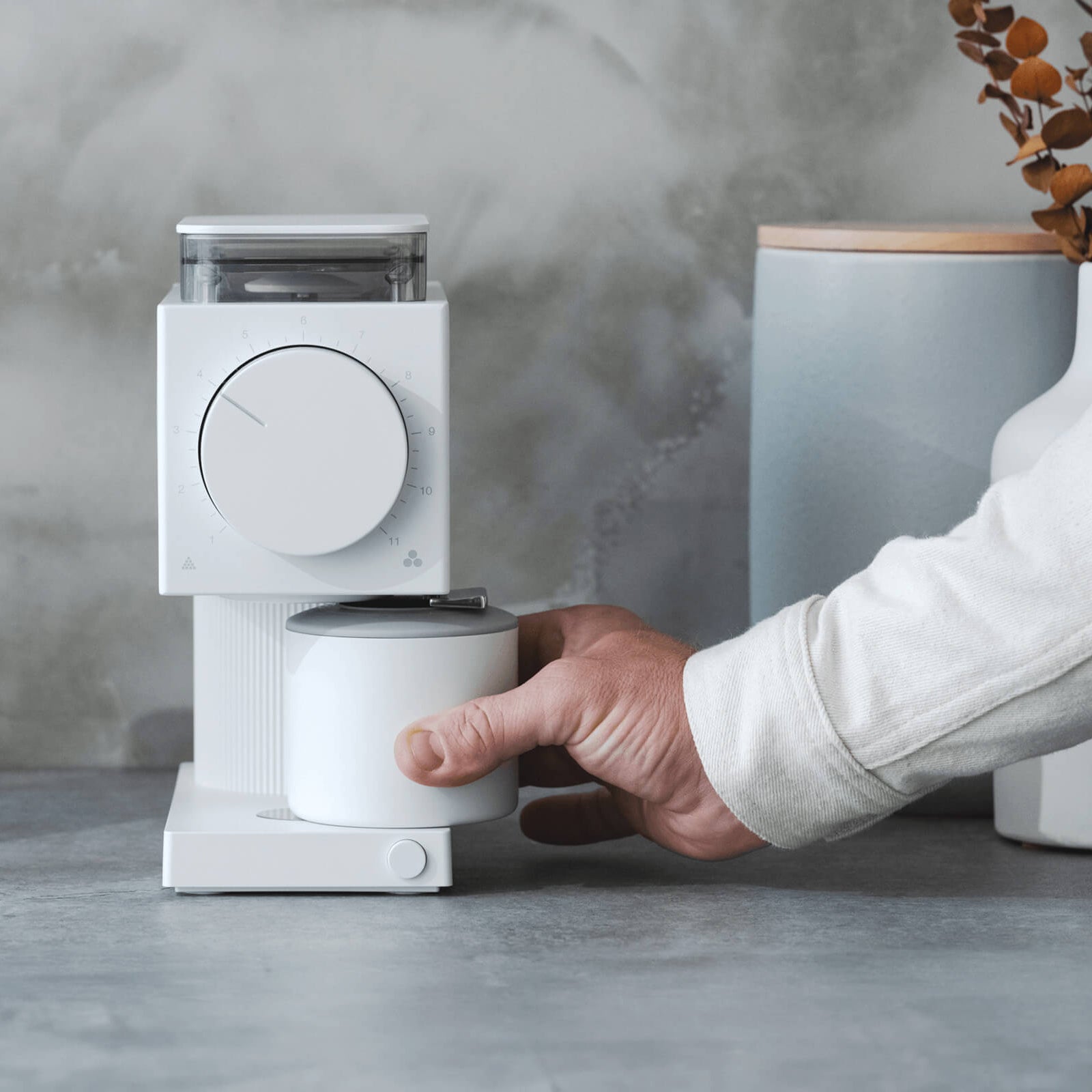 Person using a white coffee grinder with a cup on a gray surface.