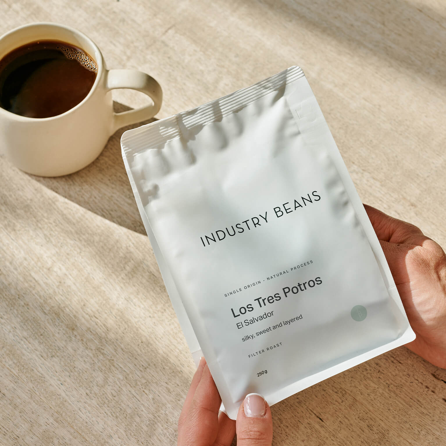 A hand holds an Industry Beans Los Tres Potros coffee bag on a light wooden table next to a cup of coffee.